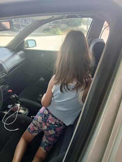 The image shows a young child, viewed from behind, sitting in the passenger seat of a car. The child has long, brown hair and is wearing a light blue ribbed tank top and floral-patterned shorts. The interior of the car is dark, with black seats and dashboard visible. A white charging cable with red beads is coiled on the passenger seat next to the child.
Through the car windows, an outdoor scene is visible. It appears to be daytime, with bright sunlight casting shadows. The exterior environment suggests a residential or urban setting, with buildings and greenery visible. A side mirror and part of the car's windshield frame are also visible, framing the view outside. The overall impression is of a casual moment, possibly during a car journey on a sunny day.