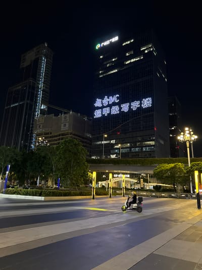 In this nighttime scene in Huangpu District, China, a modern urban environment is depicted. A tall, dark glass skyscraper dominates the left side of the frame, its facade illuminated by white neon lettering that spells out Chinese characters. Below this, a construction site is visible with a crane and scaffolding, indicating ongoing development.
To the right of the buildings, a paved pedestrian walkway features a striped pattern of light and dark gray tiles, reflecting the ambient light. Trees line the walkway, with some areas featuring green hedges. A streetlamp with multiple bulbs stands near the top of the frame, casting a warm glow.
In the middle ground, a person wearing a helmet and dark clothing is riding a scooter with a cargo box on the back. The scooter's wheels are illuminated by a yellow light. The overall atmosphere is one of urban activity at night, with a mix of established infrastructure and new construction. The lighting suggests it is a clear night.