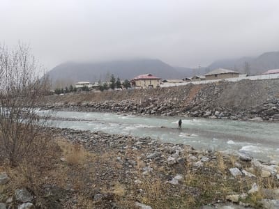 The image displays a river landscape in Qoraqiya, Uzbekistan, under an overcast sky. A wide, fast-flowing river with light-colored, turbulent water occupies the midground, characterized by numerous exposed rocks forming shallow areas and rapids. A single individual, dressed in dark clothing, stands in a rocky section of the river, holding a long, dark pole or rod, oriented towards the left. The far bank features a steep, rocky embankment, behind which multiple buildings with varied roof styles, including red-tiled and flat designs, are visible. Sparse, bare deciduous trees and a white fence line are present along this bank, with a white vehicle partially visible behind trees. Distant mountains rise under the cloudy sky, forming the backdrop. The immediate foreground and near bank consist of dry, rocky terrain with scattered bare bushes and withered vegetation.