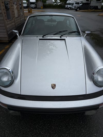The image displays a silver Porsche 911 (G-body model) parked in an asphalt lot within the City of Delta, Canada. The vehicle is positioned facing the camera, with its front hood, fenders, headlights, and bumper clearly visible. The car's surface, including the hood and fenders, is speckled with numerous water droplets, indicating recent precipitation. A Porsche crest is centrally located on the front hood. The car is equipped with round headlights featuring chrome bezels and black front bumper trim. Windshield wipers are visible at the base of the windshield. To the immediate left of the Porsche, a light-colored wooden structure with multiple rectangular windows is present, resting on a low platform. The asphalt ground shows faded yellow parking lot markings. In the background, several other vehicles are parked, including multiple recreational vehicles (RVs) and commercial vans or trucks. Specifically, two white RVs, one partially labeled "PARK" and another "ADVENTURES," are discernible, along with a dark-colored pickup truck. The sky appears overcast. No individuals are visible in the scene.