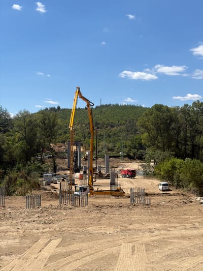 The image depicts a construction site located in the city of Kresna, Bulgaria. The scene is dominated by a large yellow construction machine with an extended boom, positioned centrally. In the foreground, several rebar cages are visible, indicating ongoing foundation work. A red dump truck and a white van are present on a dirt road in the middle ground. The background is characterized by a densely forested hillside under a bright blue sky with scattered white clouds. The overall environment suggests an outdoor construction project in a rural or semi-rural setting.