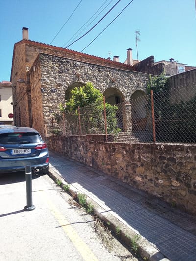 The image depicts a street scene in Castellfollit de la Roca, Spain. A dark blue Ford Fiesta, with license plate 8046 MFC, is parked on the right side of the street. The car is facing away from the camera. In the background, there is a stone building with arched openings and a red-tiled roof. A chain-link fence runs along the property line in front of the building. The street is paved with cobblestones and there is a yellow line painted on the asphalt. The sky is clear and blue. A black bollard is positioned behind the car. The scene is under bright sunlight, casting shadows on the ground.