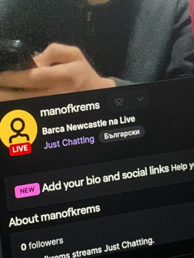 The image displays a digital screen, likely a mobile device or monitor, showing a live streaming platform interface. The screen exhibits a user profile named "manofkrems," prominently featuring a yellow circular icon with a white figure and a red "LIVE" tag. The stream's title is "Barca Newcastle na Live," categorized as "Just Chatting," and designated in "Български" (Bulgarian) language. Below this, a "NEW" prompt indicates "Add your bio and social links," followed by an "About manofkrems" section stating "0 followers." The background, blurred, shows the upper torso and hands of a person with light skin tone, wearing a dark grey or black long-sleeved garment, appearing to be observing the screen. The username "manofkrems" connects the subject to the city of Krems an der Donau, Austria. The scene depicts an individual engaging with a live stream, potentially as a streamer or viewer, in an indoor setting within Krems an der Donau, Austria.