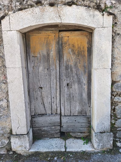 An aged, weathered double wooden door is set within a rough stone facade. The door is comprised of vertical planks with visible grain and wear, and a section of horizontal planks near the bottom. Patches of faded orange paint are present on the upper parts of the door. A small, dark keyhole is visible in the left door panel. The door is framed by an arched stone lintel and jambs made of lighter colored, roughly cut stone blocks. The surrounding wall is constructed of irregular, grey stones, with moss and some small green plants growing in crevices. The ground in front of the door is paved with stone slabs and is partially covered with dirt and dry vegetation. The scene appears to be an exterior view of an old building entrance.