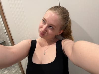 An eye-level, medium selfie shows a young Caucasian woman with blonde hair pulled back in a ponytail and light skin. She is wearing a black tank top and a silver cross necklace. Her head is tilted to the left as she looks off-camera. The background is a plain white wall with vertical grooves. The overall lighting is even and bright, with a slight shadow cast on the wall behind her head. There are no identifiable landmarks or features that would indicate a specific location within Rossiyskiy, Russia.