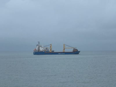 A blue-hulled cargo vessel with a white superstructure and two yellow deck cranes is centered on calm, grey-blue water. The text "BREMEN Chartering" is visible on the ship's starboard hull. The sky is uniformly light grey and overcast, contributing to a muted color palette and low visibility. No individuals or other objects are discernible in the scene. The image depicts waters near Bangor, United Kingdom.
