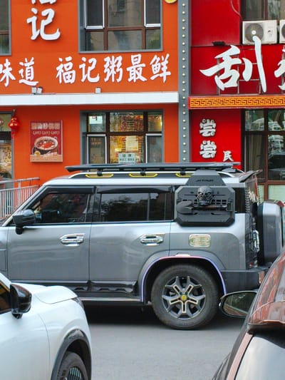 The image depicts a street scene in Jixi, China, with a focus on the facade of a building and vehicles. The building's exterior is predominantly orange and red, with large white Chinese characters adorning the upper sections. One sign on the orange section reads "味道 福记粥最鲜" which translates to "Flavor Fu Ji Zhou the Freshest". Below this, a menu displays "潮汕砂锅粥" (Chaoshan Sand Pot Congee). The red section features bold white characters, likely a restaurant name.

In the foreground, a grey SUV is parked. It has a rugged appearance with a roof rack and a distinctive black spare tire mount on the rear, which includes a skull-like design and the text "JETOUR" and "CRAZYBEE". A white car is partially visible in the lower left corner, and the side mirror of a brown car is in the lower right. The street appears to be paved. The lighting suggests it is daytime.