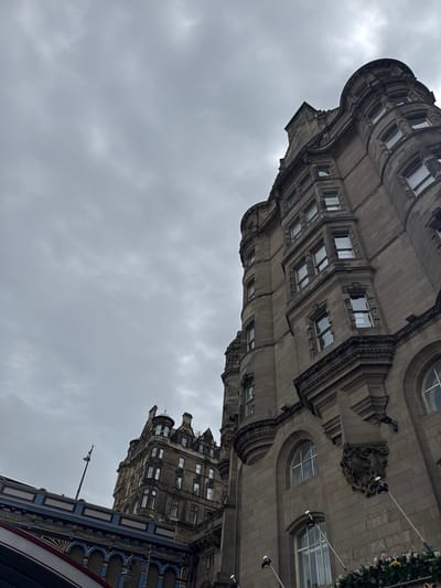 A low-angle shot captures the imposing facade of the Balmoral Hotel in Edinburgh, United Kingdom, against a heavily overcast sky. The ornate stone architecture of the hotel dominates the right side of the frame, with its distinctive curved turrets and numerous windows. To the left, a section of the elegant Edinburgh city chambers can be seen. The foreground features a portion of the North Bridge, adorned with blue and gold decorative elements and arches. A tall lamp post stands on the bridge, partially obscuring the sky. The scene is devoid of people, with the focus solely on the architectural grandeur of the city and the dramatic cloudy atmosphere.