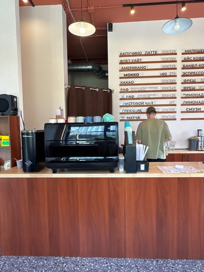 The image captures the interior of a coffee shop in Patrusheva, Russia, likely a casual establishment. In the foreground, a polished black espresso machine, possibly a Nuova Simonelli, sits on a wooden counter. To its left is a cylindrical grinder. A server, wearing a khaki t-shirt and a baseball cap, stands behind the counter, facing away from the camera, and appears to be preparing a drink. A selection of cups and utensils are visible on the counter, along with a price menu board displaying various coffee and tea options in Cyrillic script. The walls are painted a pale beige, and the ceiling features industrial lighting and exposed ductwork. A brown curtain hangs on the left side of the frame, and a shelf with items like green packaging and a yellow bag is partially visible. The flooring is a speckled gray and black tile.