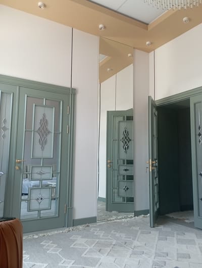 The image depicts an interior scene likely in Keles, Uzbekistan, featuring a hallway with green doors and a large mirror. On the left, a double door with glass panels and ornate decorative elements is visible. The door on the right is partially open, revealing a dark interior space. A rug with a geometric pattern covers the floor. The walls are white, and the ceiling has wooden trim with recessed lighting fixtures. The overall impression is of a modern and elegantly designed interior space.