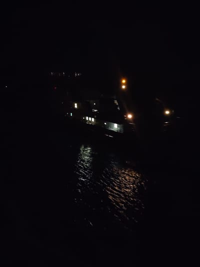 The image depicts a night scene in Gorka, Russia. A body of water reflects light from a vessel with multiple illuminated windows and running lights. A single, brighter light source is visible higher on the vessel. In the distance, faint lights suggest a city skyline. The foreground is predominantly dark, indicating the viewer is positioned at an elevated vantage point overlooking the water. The water's surface shows ripples and reflections, suggesting slight movement.