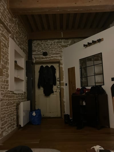 The scene appears to be an indoor renovation or construction area with exposed stone walls and flooring. A partially installed or constructed white frame structure is on the floor. Near the back, a white door hangs with coats. A blue bag sits near some stacked white panels. A shelf unit appears on the right. The location, Lyon, France, is not verifiable based on the provided imagery.