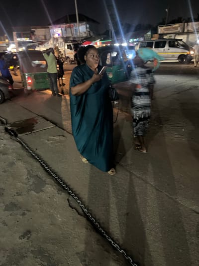 A person in a dark blue outfit stands on a street, holding a phone. Nearby, another person in a patterned dress and headscarf is blurred. Streetlights and vehicle lights illuminate the scene. There is a chain lying on the ground and vehicles are visible in the background. The photo appears to be taken at night.
