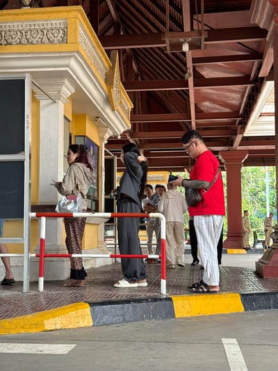 The image shows a street scene in Bavet Quarter, Cambodia. Four people are standing in a line behind a red and white barrier along a sidewalk next to a street. The first person, facing left, wears a red shirt, white striped pants, and sandals. The second person, facing left, wears a tan shirt. The third person, facing left, wears a grey suit. The fourth person, facing left, wears a tan hooded sweatshirt and leopard print pants. A yellow and black curb separates the sidewalk from the street. In the background are several people in tan uniforms. Above the people is an overhang with orange metal beams and a decorative white and yellow structure.