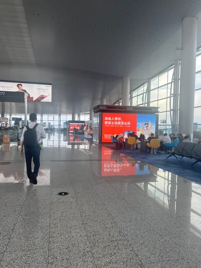 This image depicts the interior of an airport terminal in Guangzhou City, China. A man wearing a white shirt, dark pants, and a black backpack is walking away from the camera down a wide, polished floor hallway. The hallway is flanked by large windows, offering a view of the sky and possibly an airfield. In the background, there is a large digital advertisement screen displaying what appears to be travel-related content. Several people are seated in a waiting area in front of the screen, and other travelers are visible in the distance. The overall environment suggests a modern and busy transportation hub.