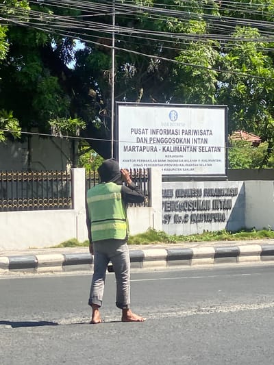 A barefoot individual wearing a reflective green vest over a dark hooded garment and grey pants stands on the right side of a paved road, facing away from the camera and towards a large sign. The sign, positioned behind a metal fence with ornate circular patterns, reads in Indonesian "PUSAT INFORMASI PARIWISATA DAN PENGGOSOKAN INTAN MARTAPURA - KALIMANTAN SELATAN" and mentions "BANK INDONESIA" at the top. The individual's right hand is raised to their head, near their ear, suggesting they might be on a phone call or adjusting their hood. Trees and power lines are visible above the sign and fence. The road has a distinct curb and a white dashed line. In the background, there is a building with a tile roof. The scene appears to be in a tropical or subtropical environment due to the lush greenery.
