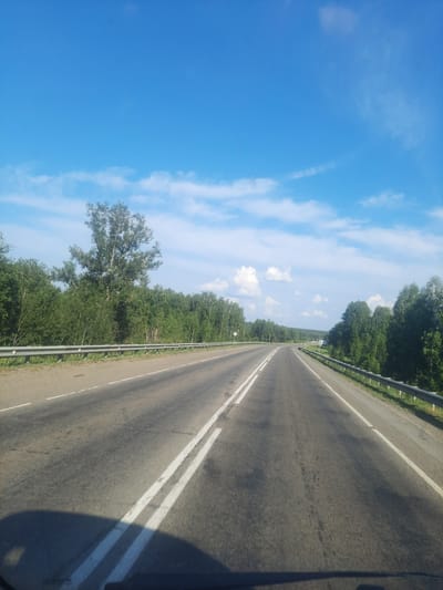 The image depicts a view of a highway in a rural setting, likely in or near Баюновские Ключи, Russia. The perspective is from a moving vehicle, looking forward along the road.
The main subjects are the road itself, flanked by lush green trees on both sides, and a bright blue sky with scattered white clouds. The road is asphalt, with distinct white lines marking the lanes. A metal guardrail runs along the right side of the road. The trees appear to be a mix of deciduous types, with dense foliage suggesting it is daytime during the warmer months.
The activity is travel, as indicated by the road and the perspective. The time of day appears to be morning or afternoon, given the bright sunlight and the clear sky with wispy clouds. The weather seems pleasant and fair. The setting is clearly rural, with the road cutting through a natural landscape dominated by trees. There are no people or specific man-made objects visible, apart from the road, guardrail, and distant, indistinct signs. The overall emotion conveyed is one of serene journeying through nature.