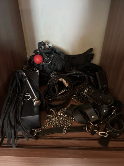 A collection of black leather restraints, including a harness, belts, wrist cuffs, a chain, and a flogger, are piled inside a wooden shelf. A red ball is attached to the harness. The background is a plain white wall. The scene is set in a closet in a residential setting.