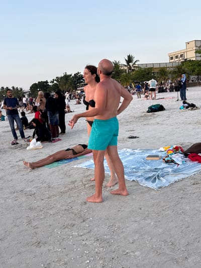 The image depicts a crowded beach scene at Cenang Beach, Malaysia, during late afternoon or early evening. In the immediate foreground, a Caucasian man with a bald head and a tanned complexion, wearing turquoise swim shorts, stands facing a Caucasian woman with dark hair, who is wearing a black bikini top and black bikini bottoms. The man has his hands on his hips, while the woman gestures with her left hand as they engage in conversation.

To their left, a person of slender build lies prone and face down on a blue towel, wearing a black bikini. Further back, numerous individuals are scattered across the sand, some standing, some seated on towels. Notable figures include a man in a dark blue shirt and jeans holding a bottle, and several women dressed in hijabs. In the midground, a light blue patterned towel is laid out on the sand, displaying a closed book and assorted clothing items. A pair of black flip-flops rests on the sand nearby. To the right midground, a woman wearing a hijab stands near a microphone and speaker setup, suggesting a performance or public address. Other individuals are gathered around this area.

The background features multiple palm trees and several multi-story buildings, including one with "OMNIA" visible on its facade, indicative of a commercial establishment like a restaurant or hotel. The sky above is a clear, light blue, suggesting the time is either approaching sunset or just after dawn. The beach sand is light-colored and shows small shell fragments and footprints.