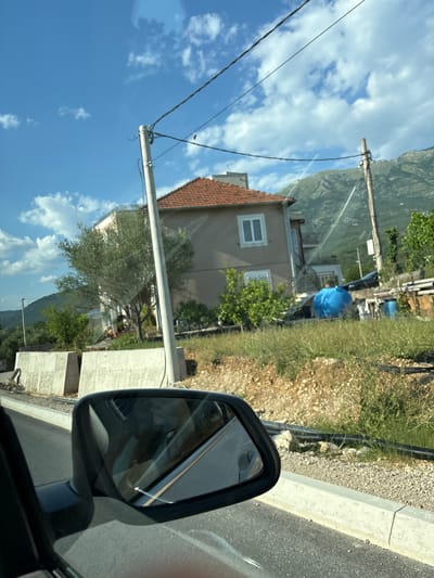This is a vertical, low-angle, outdoor, daytime shot of a residential area in Radanovići, Montenegro. The image is captured from the perspective of someone in a car, looking out the side window and towards the side-view mirror.
The scene depicts a house with a terracotta roof, beige walls, and white-framed windows. Behind the house, a steep, green mountain slopes upwards, partially obscured by trees. In the foreground, a white power pole with black wires stretches across the frame, connecting to a building on the right. Adjacent to the road, there is a grey concrete curb and a gravel area with dry grass and some green shrubbery.
The side-view mirror reflects the interior of the car, including what appears to be a person in the passenger seat wearing a white garment and a blue item. Outside the mirror's reflection, a bright blue tarp is visible, possibly covering something in the yard. The sky is a vibrant blue with scattered white clouds, suggesting a sunny day.
The overall impression is of a quiet, rural, or semi-rural setting in the Mediterranean. The lighting indicates it is likely late morning or early afternoon. There is no visible text in the image.
