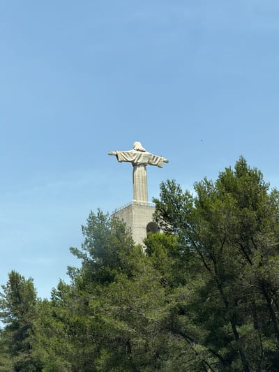 A stone statue of Jesus Christ with outstretched arms stands atop a tall, light-colored stone tower. The statue and tower are partially obscured by a dense canopy of green pine trees in the foreground. The sky is clear and blue. The statue is located at the Santuário de Cristo Rei (Sanctuary of Christ the King) in Almada, Portugal, overlooking the city and the Tagus River.