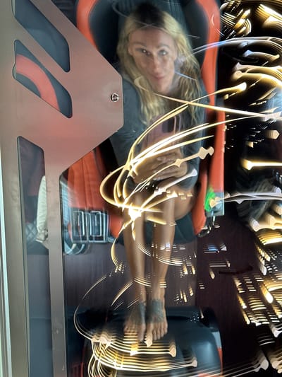 A woman with blonde hair is seated in a futuristic chair. The chair has orange and black cushioning, and a metallic frame with cutouts. The woman is wearing a dark gray shirt and dark pants. She has tattoos on her ankles. The image is distorted by light streaks, suggesting movement or a long exposure. The background is dark with more light streaks and some reflections. The location in the city of Nam Hải, Vietnam is not provided in the image or any context.