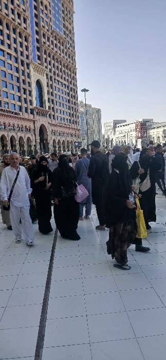 The image displays a daytime scene in a busy outdoor pedestrian area within Makkah Al Mukarramah, Saudi Arabia. A very tall, modern building with a light-colored facade, numerous rectangular windows (some reflecting blue), and multiple levels of arched architectural detailing dominates the left side. The sky above is clear and light blue. The ground consists of light-grey tiled pavement, featuring visible grout lines and a prominent darker linear expansion joint running vertically through the lower center.
A diverse crowd of individuals is walking across the tiled pavement. In the foreground-left, a man dressed in a white thobe and white pants, wearing sandals, has a dark strap bag across his chest and is walking towards the viewer's left. Several women are visible in the midground, predominantly dressed in black abayas and head coverings; one carries a purple tote bag, and another, closer to the right foreground, carries a yellow soft bag while wearing patterned pants and flip-flops. Further in the midground and background, numerous other people, mostly adults in various traditional Islamic attire, are moving in different directions. A tall, black street light with multiple fixtures is visible in the mid-distance, positioned between the prominent building on the left and other multi-story, modern buildings in the background. The overall scene depicts moderate to high pedestrian traffic in an urban environment.