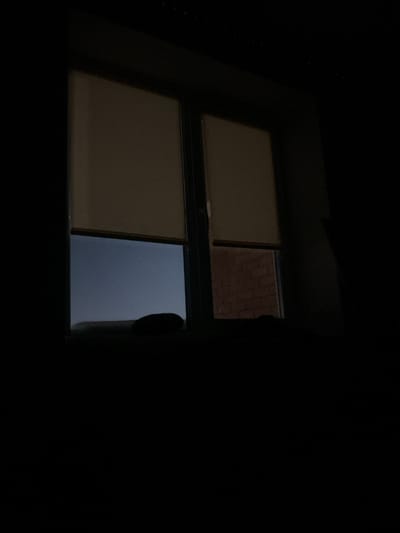 A dim, mostly dark view looking out a window. The window is split into four panes. Two panes are covered with what appear to be closed blinds, while one pane reveals a view of a brick wall and the other reflects a cloudy sky. The window frame is barely visible against the surrounding darkness, which dominates the scene.
