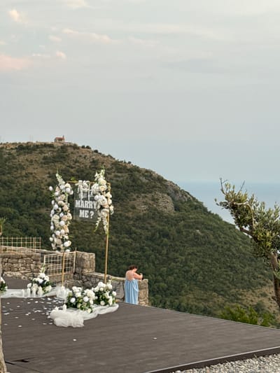 This media file depicts a romantic proposal setup in an outdoor, elevated setting overlooking a lush, green mountainside and the sea. A person in a long, light blue dress is kneeling on a dark platform, facing away from the camera and towards a decorated archway. The archway is adorned with white flowers and a sign that reads "WILL YOU MARRY ME?". White flower petals are scattered on the dark platform, and several white candles are visible near the arch.
The scene appears to be set during the day, with soft, diffused light suggesting overcast or hazy weather. The background features a steep, densely forested mountain that slopes down towards the sea, with a faint horizon line visible in the distance. An olive tree branch extends into the frame from the top left, adding to the natural aesthetic. The overall mood conveyed is one of anticipation and heartfelt emotion, likely surrounding a marriage proposal. The location context of Kamenovo, Montenegro, combined with the stunning natural scenery, suggests a picturesque and intimate proposal.