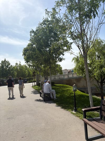 This outdoor image, taken in Kuchtumgali, Uzbekistan, captures a sunny public space featuring a paved pathway, lush greenery, and traditional architecture.
In the foreground, two individuals are walking along a wide, light-colored path. One person is dressed in a black t-shirt, light-colored pants, and dark shoes, while the other wears a light top, light pants, and sandals. Both cast clear, long shadows on the ground, indicating bright sunshine.
To the right of the pathway, a grassy bank rises, adorned with a decorative black lamp post and several dark-slatted wooden benches. Further back on one of these benches, two people are seated, one appearing to wear light clothing and another with a light blue or white head covering.
The path is bordered by numerous trees with vibrant green foliage, casting dappled shadows. In the background, traditional buildings with light-colored brickwork and ornate white decorative railings or fences are visible. A blue dome, characteristic of local architecture, rises above the buildings, suggesting a mosque or a significant cultural site. Faint text, appearing to be in Arabic script, is discernible on the building's facade.
The scene depicts a peaceful daytime environment, likely a park or garden, where people are casually strolling and resting. The weather is clear with a blue sky and some wispy clouds, contributing to the tranquil atmosphere.