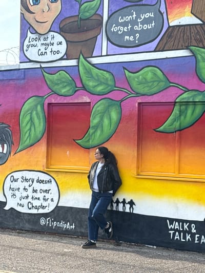 The image shows a woman standing in front of a vibrant mural. She is wearing a black jacket, a white shirt, and blue jeans, and is looking towards the left. The mural consists of comic-strip style panels with colorful backgrounds of purple, red, orange, and yellow. One panel features a plant in a pot with the speech bubble, "Look at it grow, maybe we can too," and another panel with the speech bubble, "won't you forget about me?". A plant with green leaves runs vertically along the wall. Another panel displays a speech bubble with the words "Our Story doesn't have to be over, it's just time for a new Chapter!". The mural also includes the text "@FlipadipArt" and "WALK & TALK EA" on a black background with white text, and silhouettes of people walking. The location is Gorleston-on-Sea, United Kingdom.