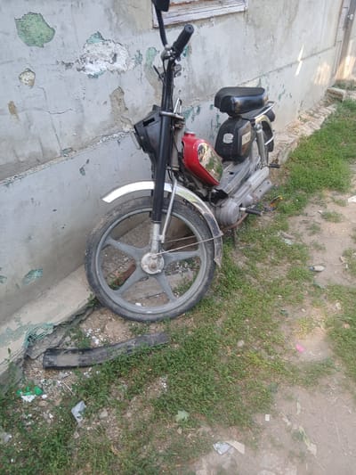 The image shows a moped lying on its side on a paved surface next to a grassy area. The moped appears to be damaged, with some parts detached and scattered around. The scene is outdoors, with natural light suggesting it is daytime. The paved area shows signs of wear and tear, with cracks and missing patches of concrete. The grassy area is overgrown with weeds and scattered with debris, including pieces of plastic and paper. The background is blurred but appears to be a residential or rural setting, possibly in Pishchanka, Ukraine, given the provided location context. There is no activity or event taking place, as the moped is stationary and appears to have been abandoned or involved in an accident. The overall impression is one of neglect and disrepair. No text is visible in the image.
