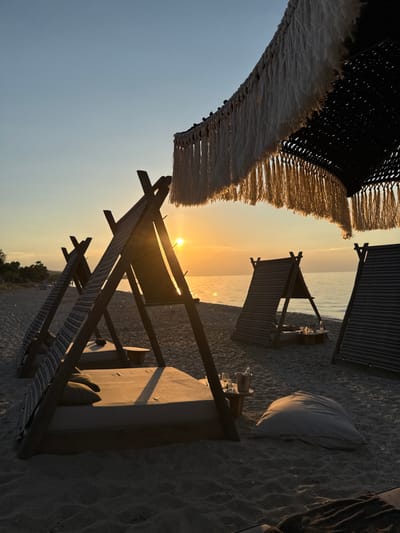 The image captures a serene beach scene at sunset in Pallini Municipal Unit, Greece. The setting is a sandy beach with several lounge areas, suggesting a resort or a well-appointed private spot. In the foreground, a large, fringed canopy, possibly part of a sunshade or tent, drapes down, its woven texture and tassels visible. Beyond this, the scene opens up to the sea, with the sun low on the horizon, casting a warm, golden glow across the water and the sky.
Several structures are arranged on the sand. One appears to be a low wooden bed or lounge with cushions and small pillows, illuminated by the setting sun. Next to it, another smaller, tent-like structure with striped fabric sides provides shade. In the distance, the calm sea stretches out, meeting a sky painted with soft hues of orange and yellow from the sunset. The overall atmosphere is peaceful and tranquil, evoking a sense of relaxation and the end of a pleasant day. The sandy ground shows distinct footprints, indicating recent activity. The time of day is clearly sunset due to the low sun and the warm lighting. The location cues like the type of beach and the general ambiance suggest a Mediterranean coastal setting, consistent with Pallini, Greece. There is no visible text in the image.