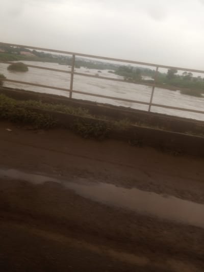 The image depicts a murky river with some vegetation and buildings in the background. A metal railing runs horizontally across the middle of the frame, separating the foreground from the river. The foreground appears to be a dirt path with some puddles of water. The sky is overcast. The scene is likely located in Nassarawa, Nigeria, as indicated by the context. There are no people visible in the image. The overall impression is one of a natural landscape under a cloudy sky.