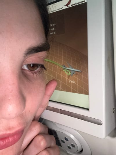 A close-up, eye-level photograph shows the right side of a person's face, including a brown eye, dark eyebrow, and portion of the nose and lips, in an aircraft cabin. The person's left hand is partially visible, holding a light gray in-flight entertainment (IFE) remote control with black buttons. Adjacent to the person's face, a rectangular IFE screen displays flight information: the "AIR INDIA" logo at the top, "powered by AIRSHOW," current altitude as "131 feet," speed as "0 mph," and temperature as "50.0 °F." The main part of the screen features a graphical map with a grid pattern, showing a grey aircraft icon with a green trajectory line, positioned directly above the text "Paris." The aircraft's reported 0 mph speed and 131 feet altitude indicate it is on the ground, specifically at Paris Charles de Gaulle Airport in Tremblay-en-France, France.