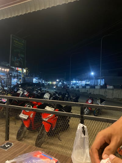 Here's a description of the image:
The photograph, taken at night in what appears to be Tanjongkapai, Indonesia, shows a street scene with parked motorcycles. The perspective is from an elevated position, possibly a balcony or upper floor of a building, looking out over the street. A metal fence or railing is in the foreground, partially obstructing the view.
Several motorcycles of varying models and colors are parked along the street. Some are oriented towards the viewer while others are positioned at an angle. License plates are visible on a few of the motorcycles, bearing the Indonesian license plate type. The vehicles seem to be closely packed together.
Across the street, there are buildings lit up with artificial lighting. A large billboard is visible on the left of the scene. It features advertising for "KUR Syariah Bank Aceh" with accompanying images and text, also showing "Apel Royal". Streetlights are present along the street, adding to the overall illumination.
In the upper right corner of the image, a person's hand is visible, holding a white plastic bag. The general lighting conditions are dim, but the light sources on the street create some contrast. The presence of overhead wires adds to the visual clutter of the scene.