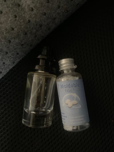 The image shows two small glass bottles on a dark, textured surface. The top bottle is clear with a silver screw-on cap and a light blue label. The label has text and an illustration of three white petals or leaves. The text on the label reads "Moldable False Teeth" and appears to indicate product details and instructions in smaller print. Inside this bottle, there are numerous small white pellets. The bottom bottle is also clear glass with a darker cap that has a spray nozzle mechanism. This bottle contains a clear liquid. The scene is dimly lit, suggesting an indoor setting or possibly nighttime. The texture of the surface suggests it might be fabric or a textured material, perhaps part of a bag or a car interior. There are no people visible in the image. The overall impression is that of manufactured products, likely related to dental care or cosmetics, being stored or transported.
