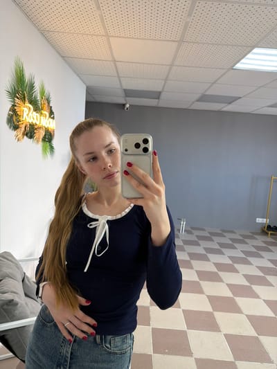A fair-skinned woman is taking a selfie in an interior space with mirrored walls. She is wearing a navy blue top with a white ribbon accent and light blue jeans. Her long blonde hair is pulled to the side. She is holding a smartphone with a silver case, visible camera lenses, and red nail polish on her fingers. The background includes ceiling panels and a checkered floor. The mirror reflects a neon sign with the Russian name "Rostova". This scene is in Berezniki, Russia.