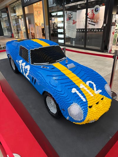 A large-scale replica of a race car, constructed entirely from blue, yellow, black, and white LEGO bricks, is displayed indoors in a commercial setting in Mont-Saint-Martin, France. The model features a predominantly blue body with a broad yellow stripe running centrally from the hood over the roof, and the number "112" is visible in white on the hood and the driver's side panel. It includes black LEGO bricks for windows and black tires with white hubs. The car is positioned on a raised, dark grey rectangular platform. A red rope barrier, supported by chrome stanchions, demarcates the display area. In the background, storefronts of retail establishments are visible through large glass windows, including a "BREAL" clothing store and another store featuring promotional signage with French text, notably "NOUVEAU & UNIQUEMENT CHEZ SEPHORA." The floor is tiled with light-colored squares, and the area is brightly illuminated by overhead lighting. No individuals are present in the immediate foreground.