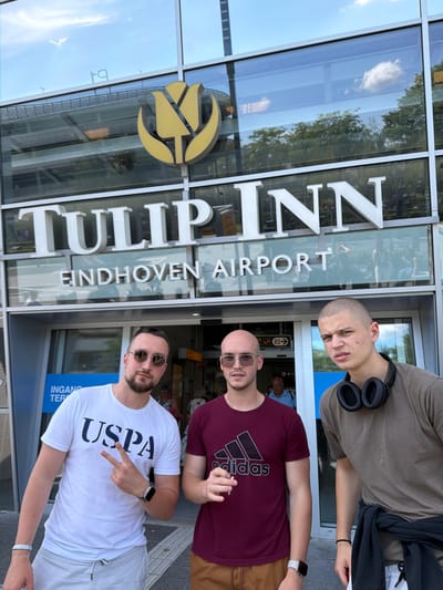 Three young men are standing in front of a building with "TULIP INN EINDHOVEN AIRPORT" signage. The man on the left wears a white t-shirt with "USPA" printed on it, sunglasses, and light-colored shorts. He is making a peace sign with his left hand. The man in the middle wears a burgundy t-shirt with the Adidas logo, sunglasses, and brown shorts. He is holding a cigarette. The man on the right wears a taupe t-shirt, has headphones around his neck, and is wearing light-colored shorts. He has a black jacket tied around his waist. The background shows the entrance of the airport building with glass windows reflecting the sky and trees.