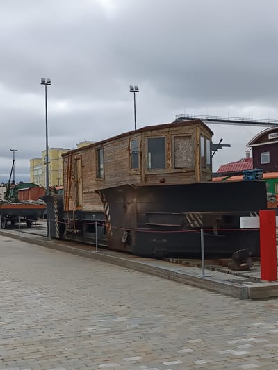 In Verkhnyaya Pyshma, Russia, a weathered, wooden train carriage sits on a flatbed train car. The carriage, with its peeling paint and boarded-up windows, appears aged and neglected. It is mounted on a dark, heavy-duty flatcar, which is itself on a railway track. Behind the carriage, a large, dark metal structure, possibly part of a snowplow or a similar piece of railway equipment, is visible. In the background, several buildings can be seen, including a tall yellow building and a smaller, darker building with a red roof. The sky is overcast, casting a muted light on the scene. The area is paved with interlocking bricks, suggesting a public space or a museum exhibit. The overall impression is of a historical artifact preserved or displayed in an outdoor setting.