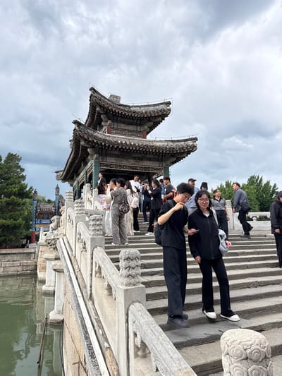 This image depicts a crowded outdoor scene at the Summer Palace (Yiheyuan) in Haidian District, Beijing, China, on an overcast day.
In the background, a large, multi-tiered traditional Chinese pavilion with dark, curved tiled roofs and red and green painted architectural elements stands prominently atop a substantial stone platform. Electrical wiring is visible near the pavilion's roofline.
A wide, grey stone bridge with numerous steps leads up to the pavilion, spanning over a body of green water visible on the left. The bridge features ornate stone balustrades with intricately carved pillars displaying cloud-scroll patterns and decorative lion-like figures at their bases. On the far left, beyond the bridge, a glimpse of a blue and red decorative archway or gateway is visible, accompanied by dense green trees.
Numerous people are present, indicating a busy tourist location. In the immediate foreground, two young adults are descending the stone steps. The male on the left, wearing dark clothing, glasses, and with dark curly hair, looks down at a smartphone held by the female. The female, on the right, also dressed in dark clothing with glasses and dark hair, carries a light-colored shoulder bag and is focused on the smartphone.
Further up the stairs and on the pavilion's lower platform, many other individuals are either ascending, descending, or standing. Several people are observed holding smartphones, actively engaged in photography. A woman in a grey patterned outfit stands near the left balustrade, taking a picture. A man in a blue shirt is walking down steps in the mid-background on the right. Another man in a grey polo shirt and black cap is visible among the crowd. A solitary figure in dark attire is discernible near the archway in the far left background.
A simple metal pipe or pole, potentially part of a utility system, is partially submerged in the green water next to the bridge, with associated cables or hoses. The sky is uniformly overcast with light grey clouds.