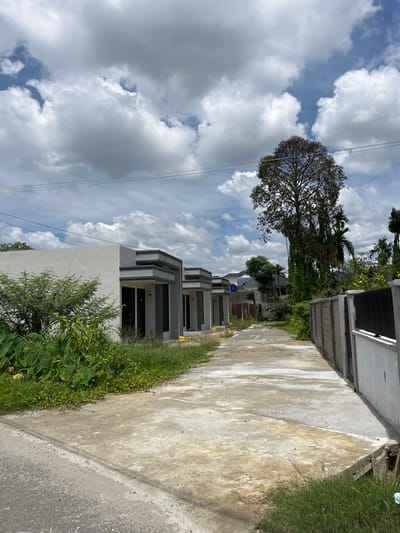 The image depicts a residential street in Langsa, Indonesia, characterized by a row of modern, minimalist houses with grey and white facades. A paved driveway runs parallel to the houses, bordered by lush green vegetation on the left and a concrete fence with a black metal gate on the right. A large tree with dense foliage stands tall near the center of the frame, contrasting with the overcast sky filled with cumulus clouds. Electrical wires are visible strung across the sky. The scene appears to be an under-construction or newly developed area, given the simplicity of the houses and the presence of some scattered yellow objects on the driveway, possibly construction materials. The environment is semi-urban, with a mix of residential structures and natural greenery.