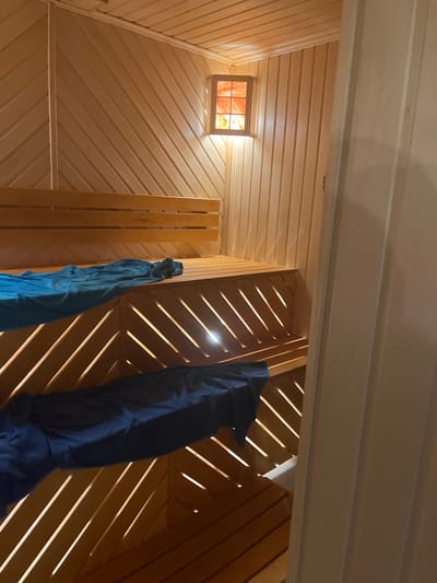 The image depicts an interior view with wooden planking on the ceiling and walls. A small, square window with a reddish glow is visible on the left wall. Two draped objects, one blue and one bluish-green, are suspended from the walls on the right side of the frame. The lighting appears to be a mix of natural light from the window and artificial light within the space. The diagonal placement of the planks on the right creates a pattern of light and shadow. The exact location in Новая Чернушка, Russia, is undeterminable from the visual information.
