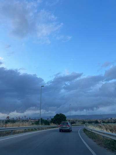A dark grey station wagon, license plate FV 564LZ, travels on a paved road curving to the right. The car is in the center of the frame, moving away from the viewer. The road is flanked by a metal guardrail on the left and right, with dry grasses and vegetation growing alongside. In the background, distant hills are visible under a dramatic sky filled with large, dark, and light grey clouds. Two lampposts are present, one on the left side of the road and another further in the distance on the right. The scene appears to be on the outskirts of Manfredonia, Italy, possibly on a highway or a provincial road leading out of the city.
