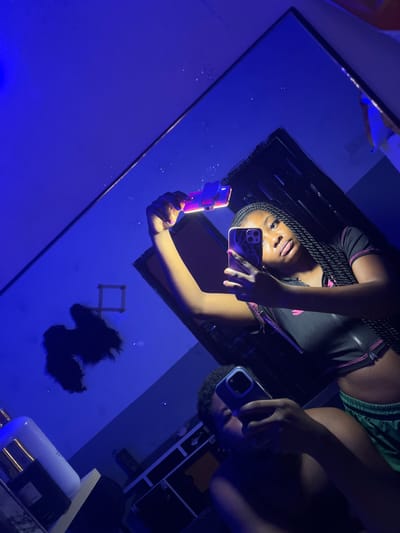 An interior scene captured as a mirror selfie in Ipetumodu, Nigeria, under dominant blue ambient lighting. The primary subject is a young adult female, reflected in the mirror, positioned centrally. She has braided hair, wears a black cropped top with pink trim, and holds a smartphone with a purple case to her face with her right hand, obscuring part of her face. Her left hand is raised, holding a second device emitting a reddish-orange glow above her head. To her lower left, a young adult male is partially reflected, holding a smartphone with a light blue case which obscures his face. He appears to wear green shorts. In the upper left background reflection, a dark, irregularly shaped object, possibly hair extensions, is visible on a blue-lit wall, below a distinct white square frame. Further left, a white bottle with a gold top rests on a surface. The overall environment is dark blue, with small reflective specks visible across the mirror surface, suggesting dust or decorative elements.