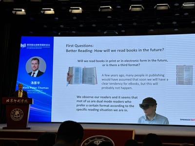 The image captures a presentation in a conference hall, likely within the Daxing District of China, given the "北京印刷学院" (Beijing Institute of Graphic Communication) branding visible on the podium and in the background. A speaker, identified as Niels Peter Thomas, Executive Officer of BMJ Publishing, stands at a wooden podium with the institute's emblem. He is addressing an audience, though they are mostly out of frame.

The main focus is a large projection screen displaying presentation content. The screen features the title "First Questions: Better Reading: How will we read books in the future?". Below this, text discusses reading habits, mentioning print versus electronic forms and a potential third format. Images on the screen include a hand holding an open book, a close-up of a book's pages, and a diagram of a book. There is also an image of a man wearing a VR headset, illustrating a point about future reading experiences. The overall ambiance is that of an academic or industry conference.