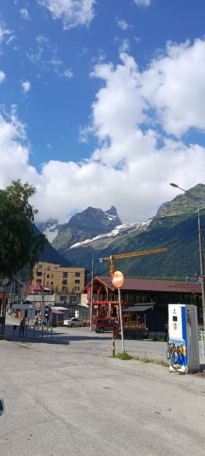 The image captures a scenic view of Dombay, Russia, with a focus on a street scene set against a backdrop of majestic mountains. The sky is a vibrant blue, dotted with fluffy white clouds. In the foreground, a paved road leads into the town, with several vehicles parked and in motion. To the right, a modern electric vehicle charging station stands prominently, featuring blue and white panels and coiled charging cables. A red SUV is parked to its left.

The town itself is characterized by a mix of architectural styles. A multi-story building with a yellow facade, featuring numerous windows and balconies, is visible in the mid-ground. Adjacent to it, a rustic wooden structure with a red roof appears to be a restaurant or cafe, with outdoor seating. A prominent yellow construction crane looms behind these buildings, suggesting ongoing development.

Further into the background, the dramatic slopes of the Dombay mountains rise, their peaks partially obscured by clouds and snow. Lush green trees and vegetation cover the lower parts of the mountains. A single street lamp with a modern design stands on the right side of the image, illuminating the street. A circular traffic sign with a red horizontal bar indicates "no entry" or a similar restriction. A few individuals are present on the street, some walking and others near shops, contributing to the sense of a bustling, albeit picturesque, town. The overall impression is one of a vibrant mountain resort town, blending natural beauty with modern amenities.