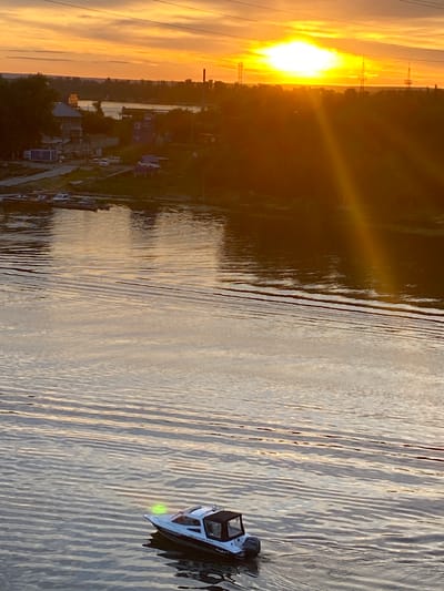 The image depicts a sunset scene over a body of water, likely a river, with a small boat visible on the right. The sun, a bright orb casting a golden glow, is low on the horizon, illuminating the sky with warm oranges and yellows. Power lines are silhouetted against the bright sky, suggesting an industrial or semi-urban setting. On the left bank, there are buildings and trees, with some structures appearing to be houses or small businesses. The water is calm, with gentle ripples reflecting the light from the sky. The boat, a white vessel with a dark roof, is moving away from the viewer, its bow pointing towards the right side of the frame. A green light is visible on the front of the boat. The overall mood is serene and peaceful, capturing the beauty of a sunset. The location is identified as Novokuybyshevsk, Russia. The time of day is sunset. There is no visible text in the image.