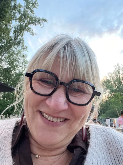 The image is a close-up selfie of an adult female with blonde and grey hair and fair skin, captured outdoors in Veauchette, France. She is wearing black, angular eyeglasses and a light-colored knitted cardigan over a dark brown collared shirt. A thin gold-colored necklace with a small, circular pendant adorns her neck, and reddish-maroon drop earrings are visible. The subject is smiling, revealing her teeth and pronounced crow's feet around her eyes. The background features a blue sky with scattered clouds and lush green trees, indicating an outdoor setting during late afternoon or early evening. A dark umbrella or canopy is partially visible on the left. Distant, out-of-focus elements on the right include other individuals, tables, and a white cup, suggesting an outdoor restaurant or gathering space.